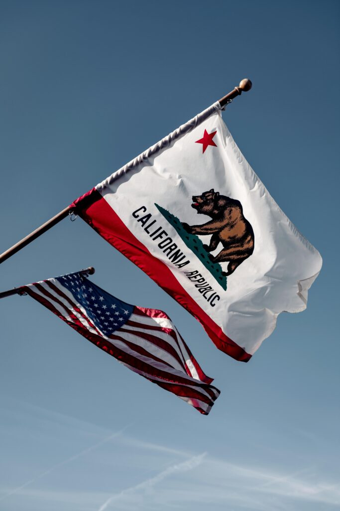 Two flags wave against a clear blue sky: the California state flag, featuring a bear, and the United States flag below. The scene conveys pride and unity.