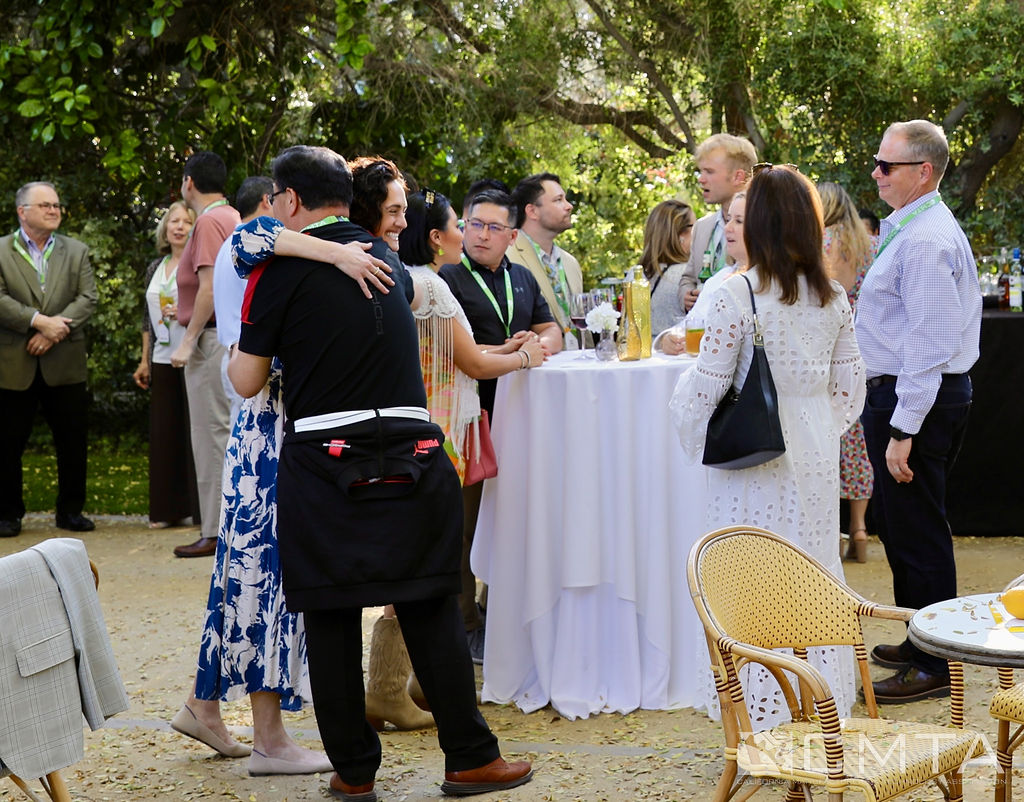 A group of people gathers outdoors at a social event, smiling and chatting beside a white table with drinks. Trees and sunshine create a warm, inviting atmosphere.