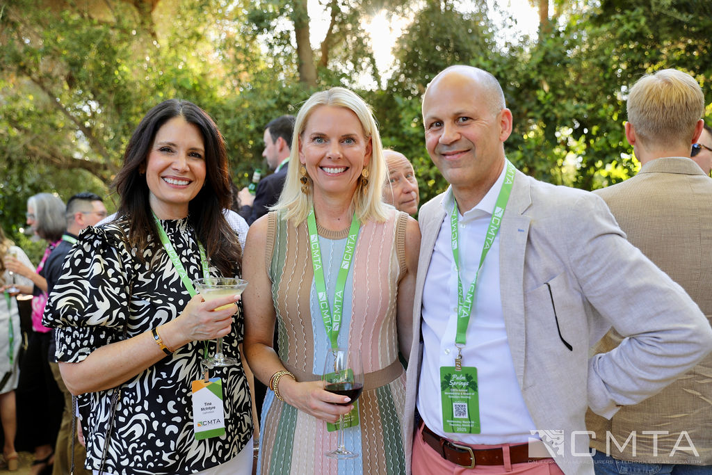 Three people smiling at an outdoor event, wearing casual business attire with event lanyards. They hold drinks, surrounded by trees and attendees.