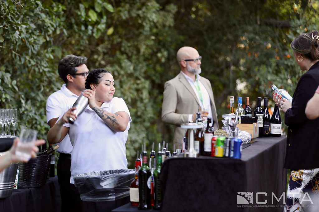 Two bartenders in white shirts prepare drinks at an outdoor event, shaking a cocktail mixer. A man in a suit and a woman are watching, with bottles and glasses on the table. The setting is casual and lively.