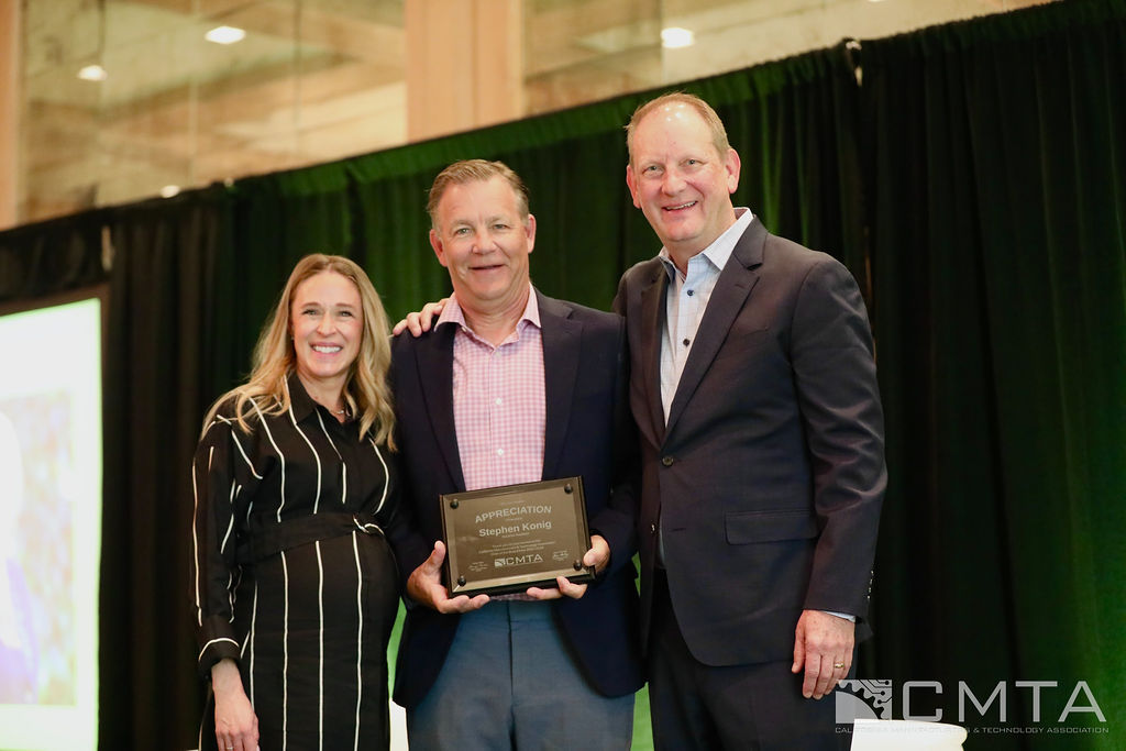 Three people stand smiling on a stage. One holds a plaque reading "Appreciation." The backdrop features a green curtain and a logo.