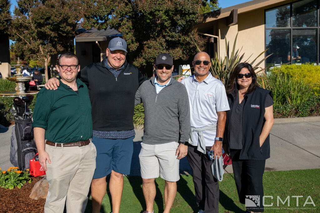 A group of five people smiling outdoors on a sunny day, standing on a grassy area near a building. The scene conveys a sense of camaraderie and joy.