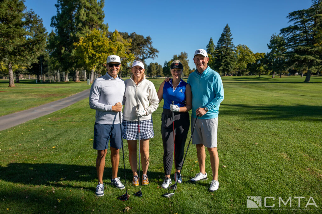 Four people stand on a sunny golf course, smiling and holding clubs. They wear casual golf attire, surrounded by green grass and trees. Logos are visible.