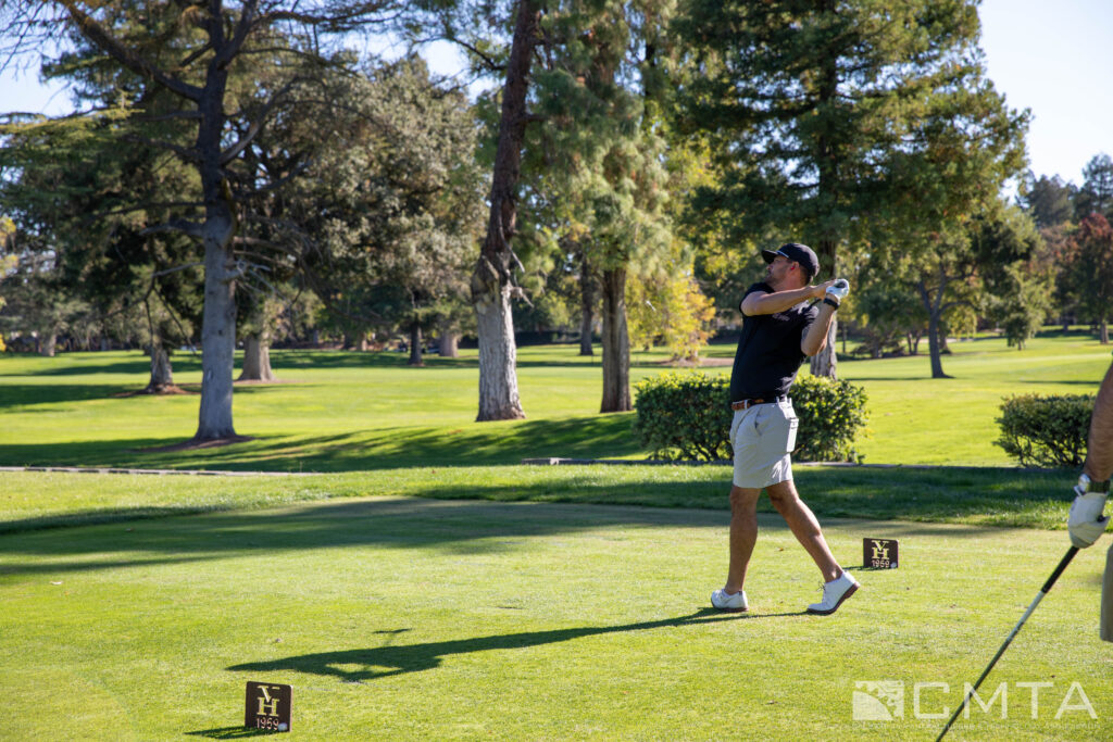 A golfer in mid-swing on a sunny course, surrounded by lush green trees. The relaxed and focused atmosphere highlights the enjoyment of the game.