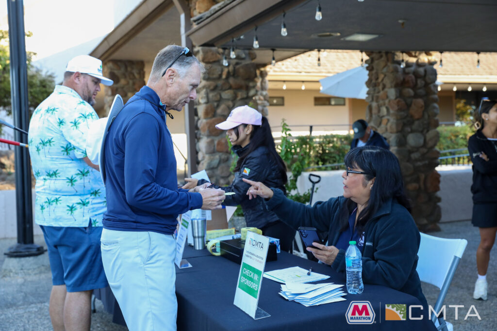 A man signs in at a check-in table outdoors, interacting with a seated woman. Another man in a printed shirt checks in nearby. A sign reads, "CHECK-IN SPONSORS."