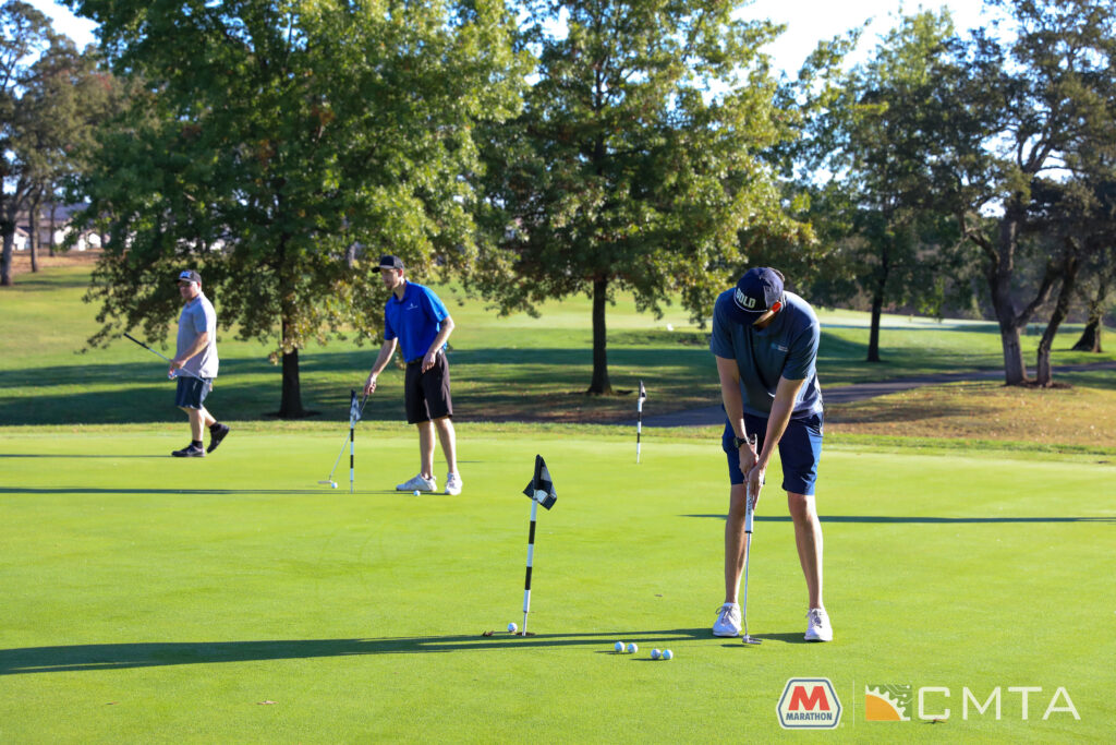 A golfer in a blue shirt and cap focuses intently as he lines up a putt on a bright green course, with trees in the background. Two other golfers are in the distance. Logos for Marathon and CMTA are visible in the corner.