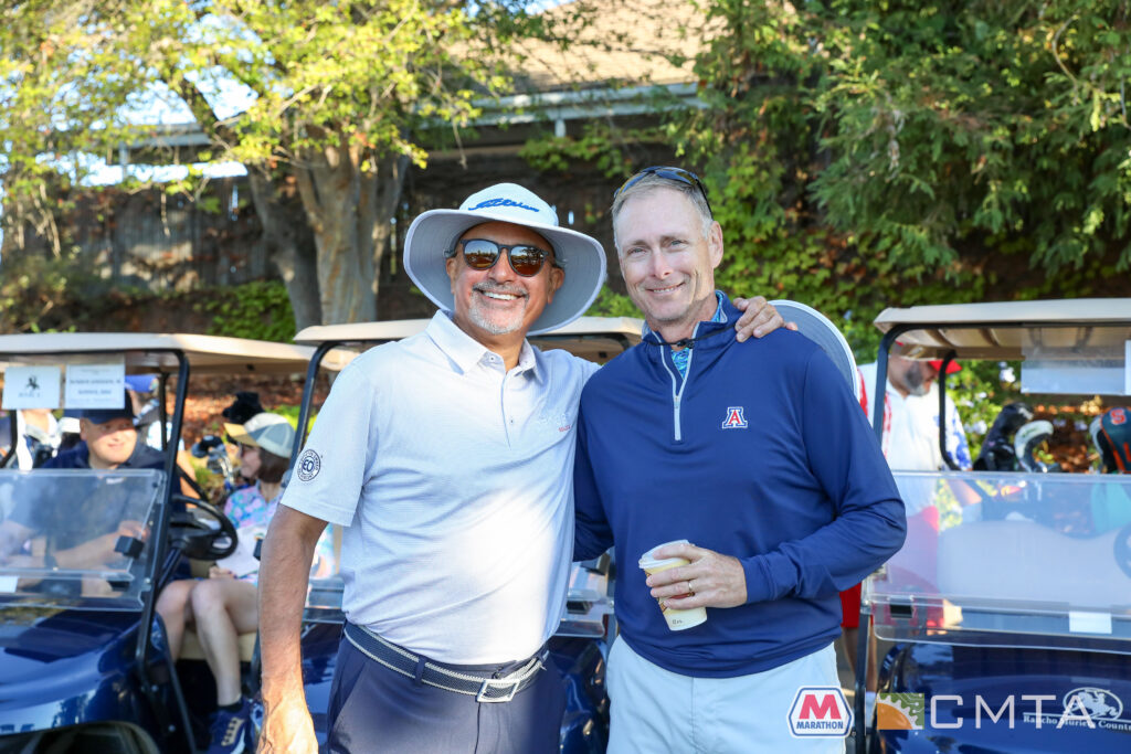 Two men smiling, standing next to each other on a sunny day at a golf course with golf carts in the background. Both are dressed in casual golf attire.