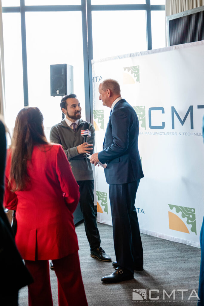 A man in a suit is being interviewed by another man holding a microphone. A woman in a red suit stands nearby. A "CMTA" banner is in the background. The setting appears professional and formal.