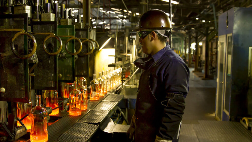 A worker in protective gear and a helmet oversees glowing orange glass bottles on a conveyor in a dimly lit factory, conveying focus and industrial intensity.